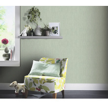 Woonkamer met lichtgroen behang met fijne structuur, een fauteuil met bladpatroon en decoratieve planten op een wandplank.