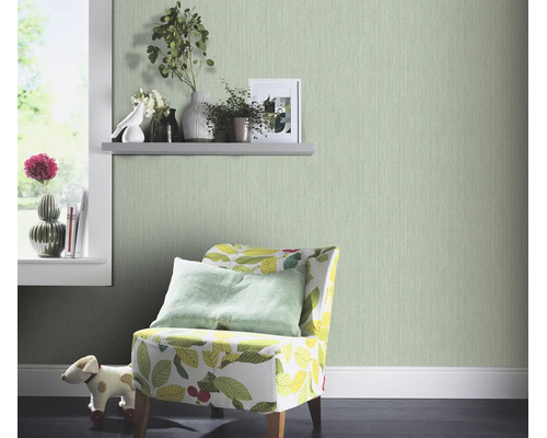 Woonkamer met lichtgroen behang met fijne structuur, een fauteuil met bladpatroon en decoratieve planten op een wandplank.