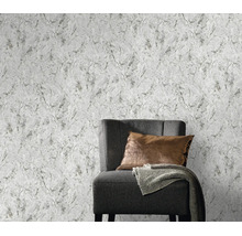 Vliesbehang met gestructureerde betonlook achter een moderne fauteuil met kussen en plaid.