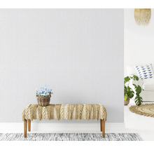 Gestructureerd vliesbehang op een muur in een lichte woonkamer met een bankje met franjes en een plant in een mand.