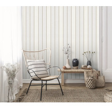 Een vliesbehang met verticale strepen in een licht ingerichte woonkamer met een rotan stoel en een houten bankje.