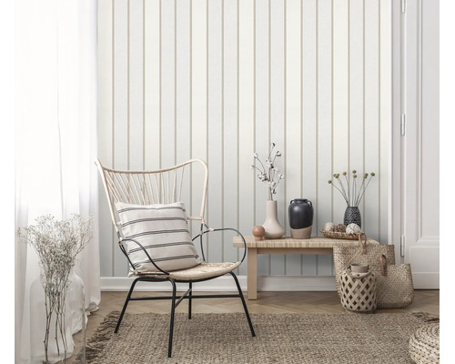 Een vliesbehang met verticale strepen in een licht ingerichte woonkamer met een rotan stoel en een houten bankje.
