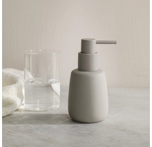 Zeepdispenser in mat grijs naast een drinkglas en een handdoek op een licht oppervlak.