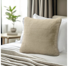 Zacht sierkussen van imitatiebont in beige op een bed met wit beddengoed, op de achtergrond een bijzetafel met plant.