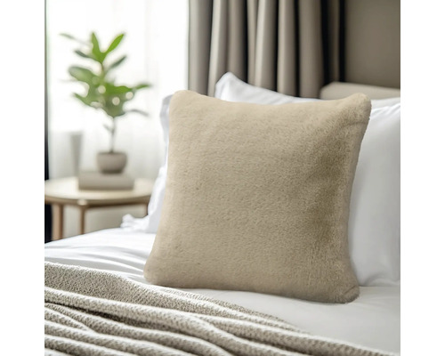 Zacht sierkussen van imitatiebont in beige op een bed met wit beddengoed, op de achtergrond een bijzetafel met plant.