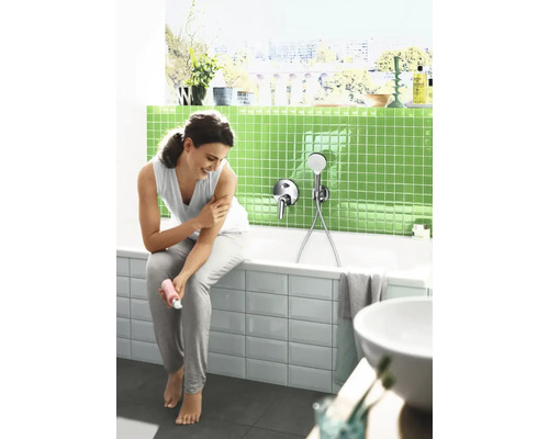 Vrouw zit op de rand van een bad in een moderne badkamer met groene mozaïektegels en een badmengkraan met handdouche.