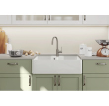 Witte keramische boerenwastafel met moderne keukenkraan in een lichte keuken met groene kasten.