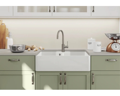 Witte keramische boerenwastafel met moderne keukenkraan in een lichte keuken met groene kasten.