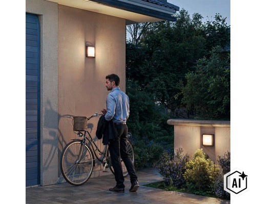 Een man met een fiets voor een huis bij avondschemering, verlicht door twee moderne vierkante buitenwandlampen aan de gevel. (Image AI modified or generated)