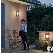 Een man met een fiets voor een huis bij avondschemering, verlicht door twee moderne vierkante buitenwandlampen aan de gevel. (Image AI modified or generated)