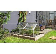 Een kleine koude bak van glas met aluminium frame staat op een stenen rand in een tuin.