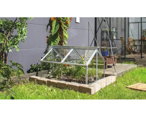 Een kleine koude bak van glas met aluminium frame staat op een stenen rand in een tuin.