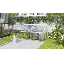 Een kleine koude bak van aluminium en transparante platen staat in een verzorgde tuin voor een muur met planten.