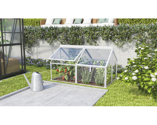 Een kleine koude bak van aluminium en transparante platen staat in een verzorgde tuin voor een muur met planten.