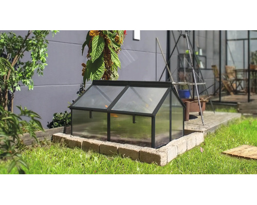 Een kleine koude bak van metaal met lichtdoorlatende platen, geplaatst op een stenen sokkel in een tuin.