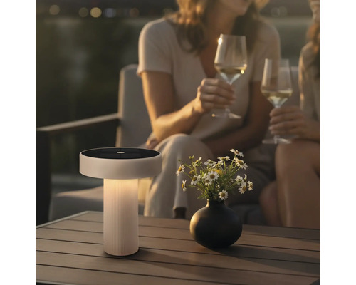 Moderne solartafellamp met geïntegreerd zonnepaneel op een terrastafel naast een bloemenvaas bij twee personen met wijnglazen.