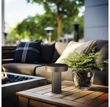 Moderne ledtafellamp voor buiten op een houten tafel naast een plant en een vuurkorf op een terras.