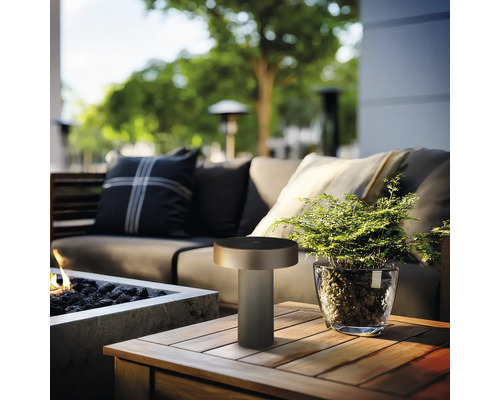 Moderne ledtafellamp voor buiten op een houten tafel naast een plant en een vuurkorf op een terras.