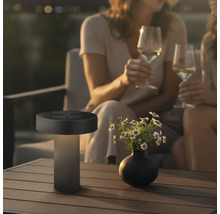 Moderne led tafellamp voor buiten met zonnepaneel op een tuintafel naast een bloemenvaas en personen op de achtergrond