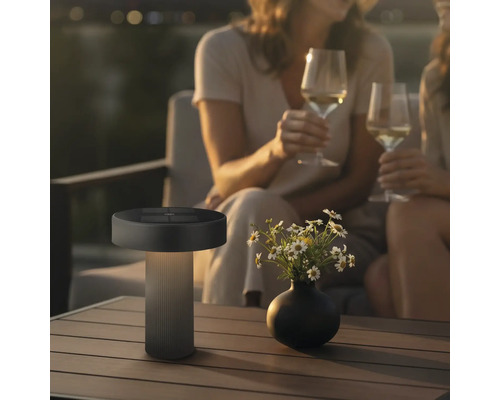 Moderne led tafellamp voor buiten met zonnepaneel op een tuintafel naast een bloemenvaas en personen op de achtergrond
