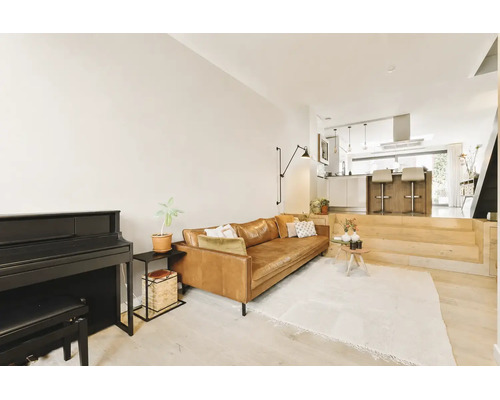 Open woonruimte met lederen bank, piano en verhoogd keukengedeelte met kookeiland in een modern en licht interieur.