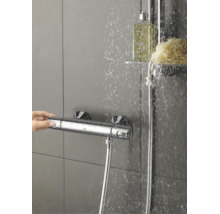 Een Grohe douchethermostaatkraan op een betegelde wand met stromend water en een hand die de knop bedient.