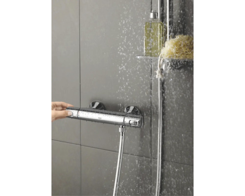Een Grohe douchethermostaatkraan op een betegelde wand met stromend water en een hand die de knop bedient.