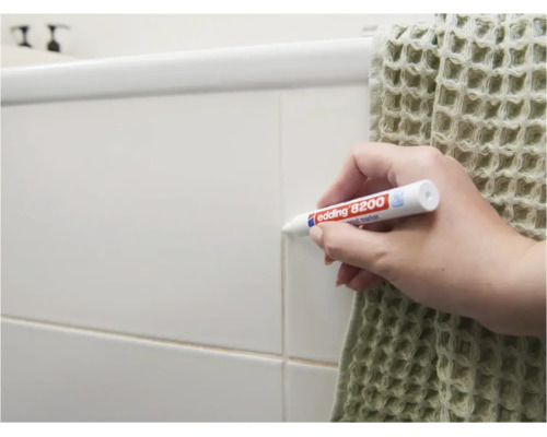 Persoon gebruikt een Edding 8200 voegenstift om de voegen van tegels aan een muur in een badkamer op te frissen.