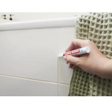 Een hand gebruikt een Edding 8200 voegenstift om tegelvoegen op een muur in de badkamer bij te werken.