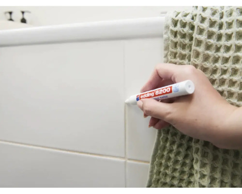 Een hand gebruikt een Edding 8200 voegenstift om tegelvoegen op een muur in de badkamer bij te werken.