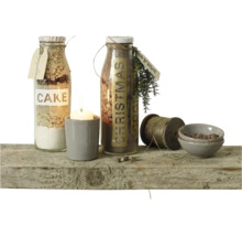 Twee glazen flessen met bakmixen, een kaars in een beker, een garenklos en kleine schaaltjes op een rustieke houten plank ter decoratie.