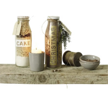 Twee glazen flessen met bakmixen, een kaars in een beker, een klos garen en twee kleine schaaltjes op een rustieke houten plank.