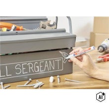 Een hand schrijft op een grijze metalen gereedschapskist met een Edding 750 lakmarker op een houten tafel. (Image AI modified or generated)