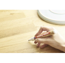 Een hand gebruikt een Edding 8900 meubelstift om krassen op een houten oppervlak bij te werken.