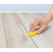 Een hand gebruikt een klein geel blokje om een voeg tussen de planken van een lichte houten laminaatvloer te reinigen.