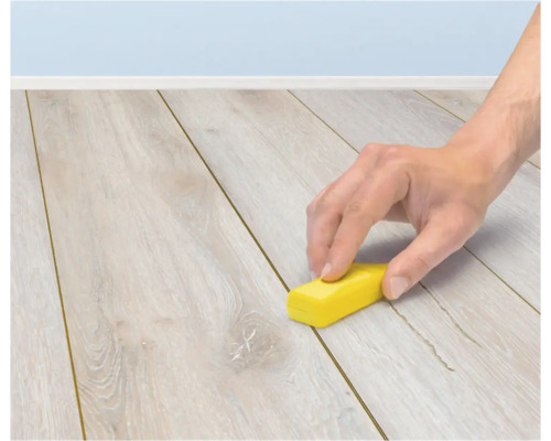 Een hand gebruikt een klein geel blokje om een voeg tussen de planken van een lichte houten laminaatvloer te reinigen.