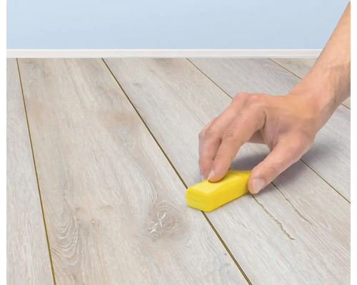 Een hand gebruikt een reparatiewasstift om krassen op een laminaatvloer te herstellen.