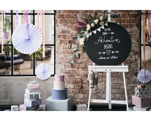 Een rond krijtbord met tekst op een schildersezel, omringd door stapels cadeaus, papieren waaiers en bloemen voor een bakstenen muur.