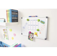 Wit whiteboard met aluminium frame, voorzien van een weekplanner, gemonteerd aan een muur naast een boekenplank met leerboeken.