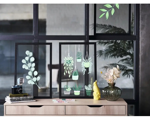 Twee zwarte glazen lijsten met plantentekeningen op een licht houten dressoir met decoratieve objecten voor een glazen wand.