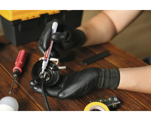 Een persoon met werkhandschoenen markeert een elektrisch onderdeel met een Edding lakmarker op een houten tafel.