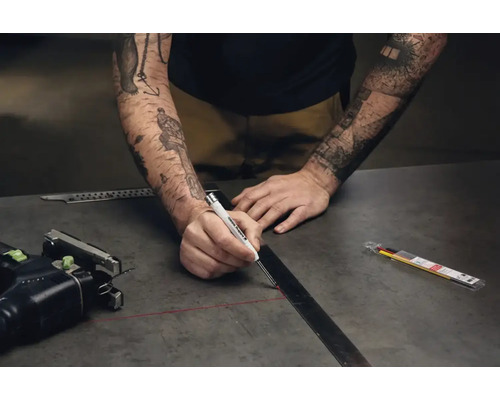 Een persoon tekent met een Edding diepgatmarker een lijn langs een metalen liniaal op een donker werkblad.