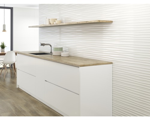 Keukenscène met witte onderkasten, een houten plank en een muur met een golvende tegelstructuur.
