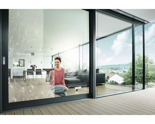 Vrouw maakt ramen schoon in een moderne woonkamer met glazen schuifdeuren en terras.