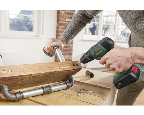 Een persoon boort met een accuschroevendraaier een gat in een houten plank met pijpverbindingen.