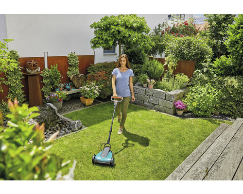 Een vrouw gebruikt een gazonmaaier in een verzorgde tuin met rotstuin en weelderige beplanting.