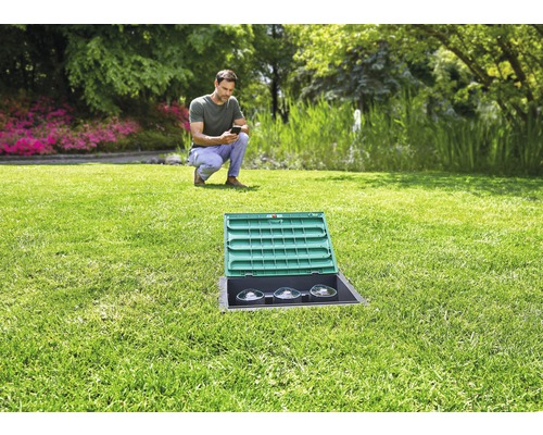 Tuinlandschap met een man die een smartphone bedient, op de voorgrond een in het gazon ingebouwde stroomverdeler met open deksel.