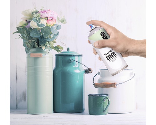 Decoratieve scène met bloemen, gekleurde melkkannen en een hand die een spuitbus Maston One vasthoudt.