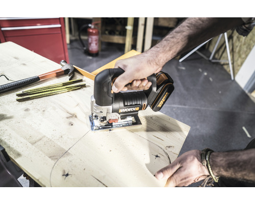 Een persoon zaagt een houten plank met een Worx decoupeerzaag. Gereedschappen zoals een hamer en een duimstok liggen klaar.