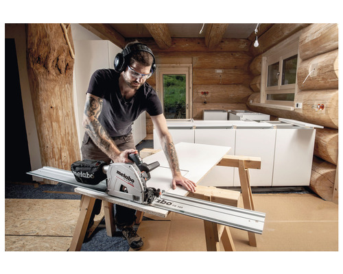 Een vakman met veiligheidsbril en gehoorbescherming zaagt een plaat met een Metabo invalzaag in een ruimte met houten wanden.